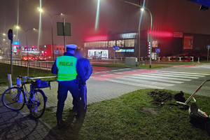 Na zdjęciu widać policjanta, miejsce zdarzenia drogowego, uszkodzony rower oraz bariery drogowe. W tle widać przejście dla pieszych i przejazd dla rowerzystów.