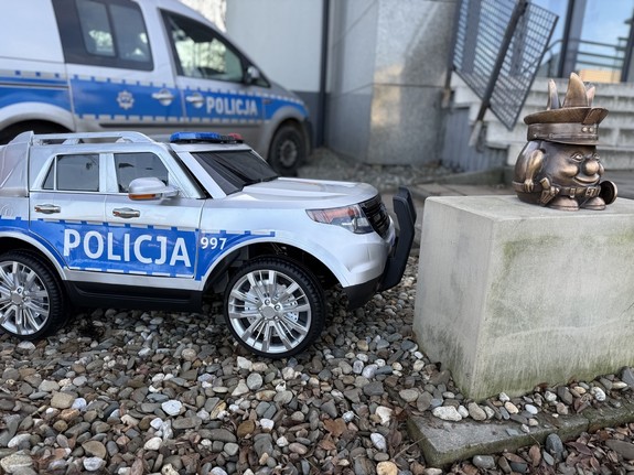 Na zdjęciu widać Żorka Policjanta, samochodzik akumulatorowy, a w tle komendę oraz radiowóz.