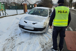 Na zdjęciu widać jasny samochód pokryty śniegiem, policjanta oraz drogę.