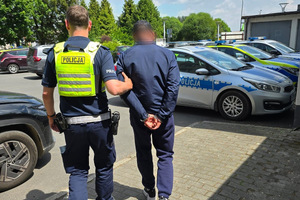 Na zdjęciu widać policjanta, który prowadzi zatrzymanego mężczyznę. W tle widać radiowozy.