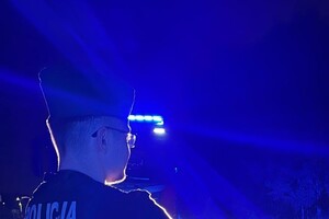 Na zdjęciu widać policjanta oraz radiowóz z włączonymi sygnałami świetlnymi w porze nocnej.