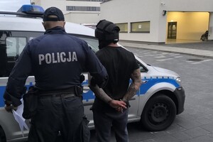 Na zdjęciu widać policjanta, radiowóz i zatrzymanego mężczyznę w kasku ochronnym i kajdankach.
