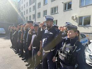 Na zdjęciu widać policjantów i policjantki żorskiej komendy Policji zgromadzonych na parkingu przed budynkiem i oddający honor poległemu koledze.