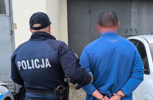 Na zdjęciu widać radiowóz, policjanta i zatrzymanego mężczyznę w kajdankach.